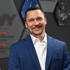 Smiling man in a blue suit and white shirt stands in front of a gray wall with geometric designs.