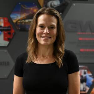 Smiling woman with brown hair in a black shirt stands in front of a dark, modern wall with hexagon images.